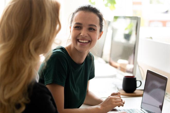 Woman leaning forward toward her laptop and smiling at another woman whose back is toward us