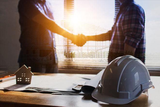 Two people shaking hands in the background, paperwork and a safety helmet on the desk