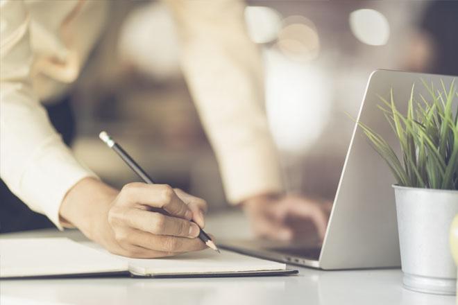 woman's hand writing on a notebook with a pencil while the woman leans toward a laptop and supports herself with her other hand